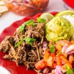 Slow cooker barbacoa served in soft corn tortillas with guacamole, pico de gallo, and lime on a red plate.