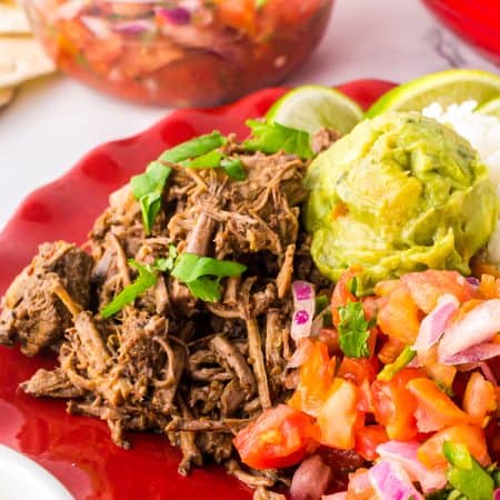 Slow cooker barbacoa served in soft corn tortillas with guacamole, pico de gallo, and lime on a red plate.