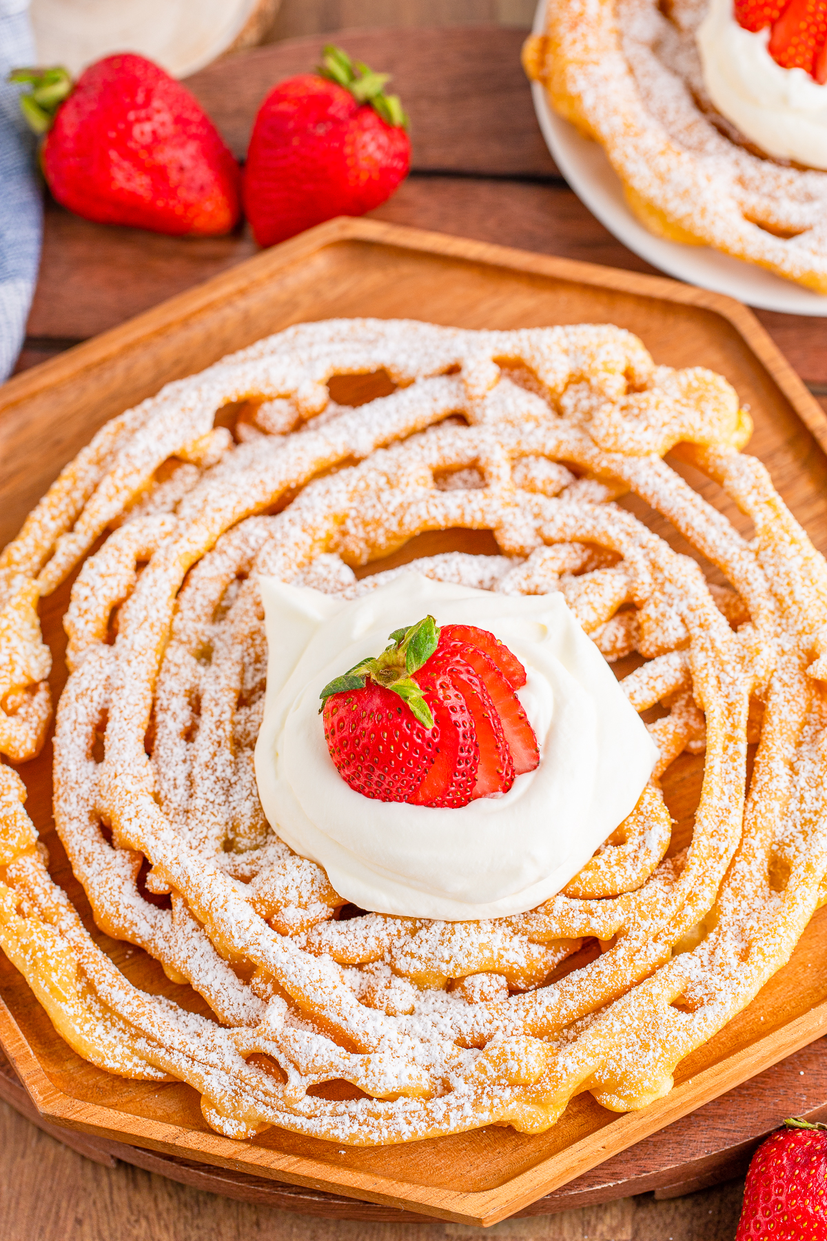 Overhead of one Funnel Cake on wooden plate topped with powdered sugar, whipped cream and strawberries.
