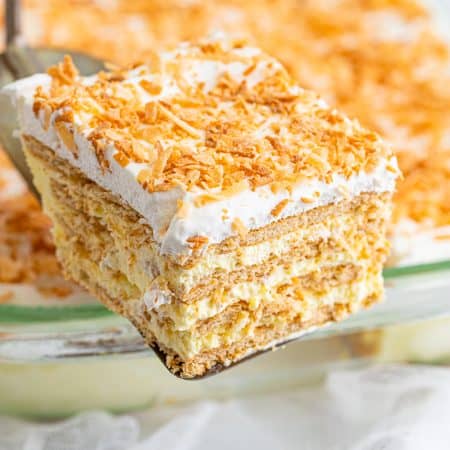 A slice of icebox cake with toasted coconut topping being lifted from the baking dish showing creamy layers.