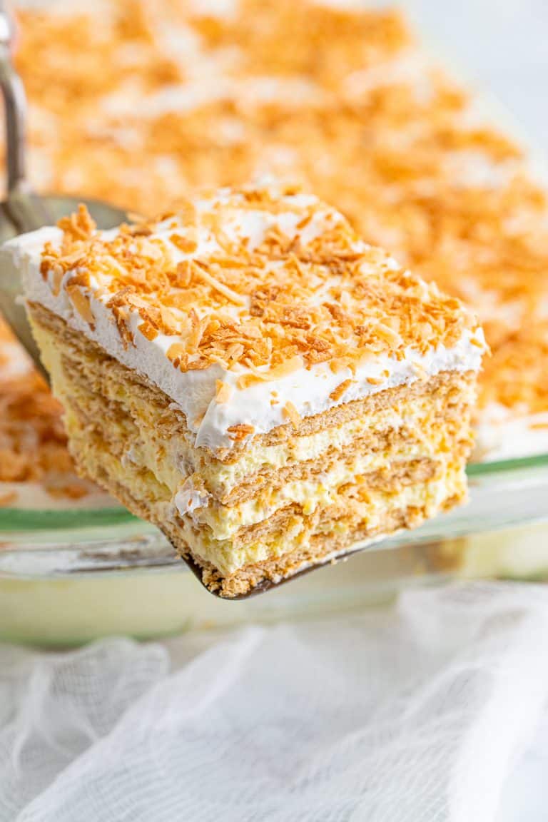 A slice of icebox cake with toasted coconut topping being lifted from the baking dish showing creamy layers.