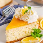 Slice of Lemon Cheesecake topped with whipped cream, lemon and mint on white plate.