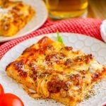 Slice of Sheet Pan pizza on white plate with glass of beer in background.