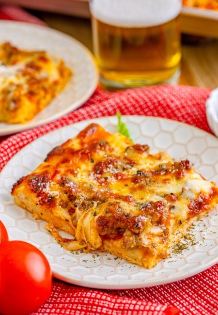 Slice of Sheet Pan pizza on white plate with glass of beer in background.