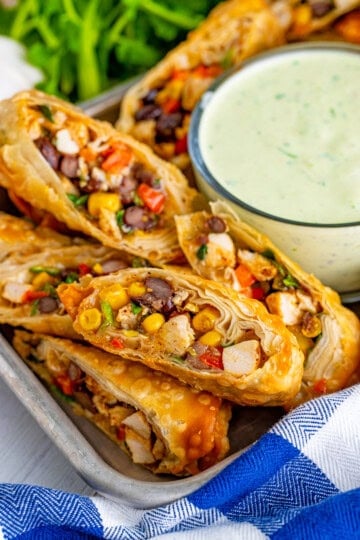 Crispy Copycat Chilis Southwest Egg Rolls cut diagonally in a metal serving tray with a bowl of avocado ranch dipping sauce, fresh cilantro, and a blue and white striped towel.