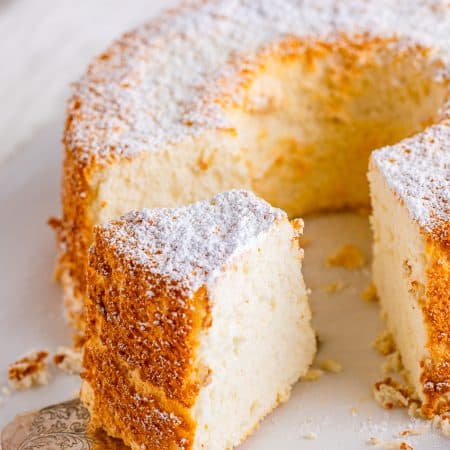 A whole angel food cake dusted with powdered sugar on a cake stand with one slice removed