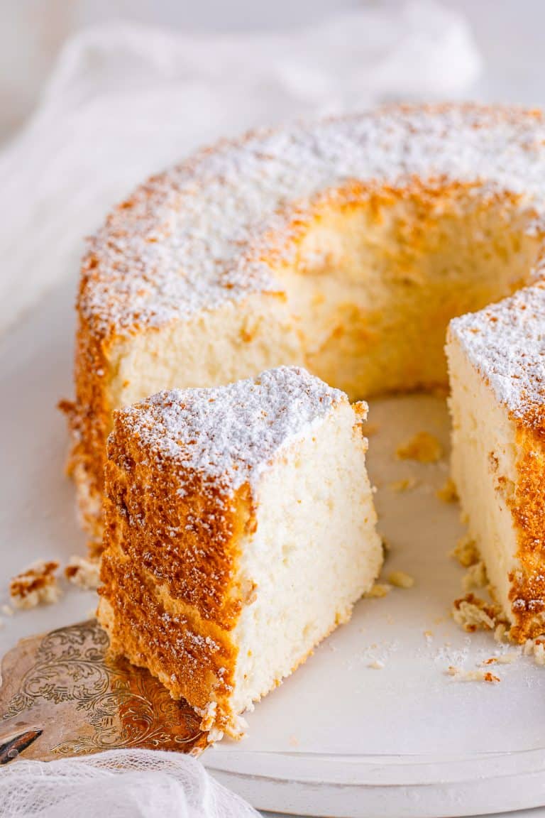A whole angel food cake dusted with powdered sugar on a cake stand with one slice removed