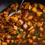 A skillet of glossy bourbon chicken in a rich brown sauce garnished with sliced scallions