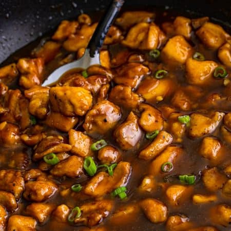 A skillet of glossy bourbon chicken in a rich brown sauce garnished with sliced scallions