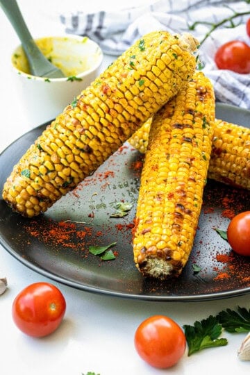 Charred grilled corn on the cob brushed with herb butter on a dark plate.