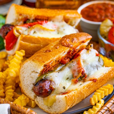 Two Grilled Italian Sausage Sandwiches on wicker board surrounded by french fries.