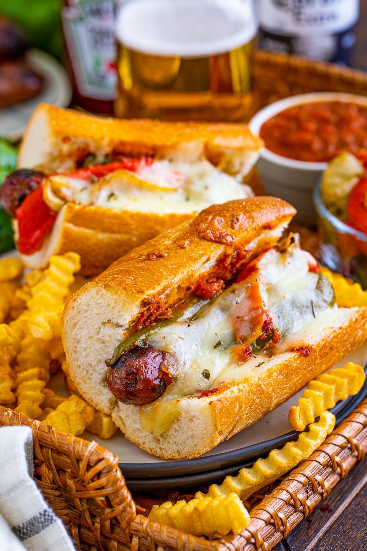 Two Grilled Italian Sausage Sandwiches on wicker board surrounded by french fries.