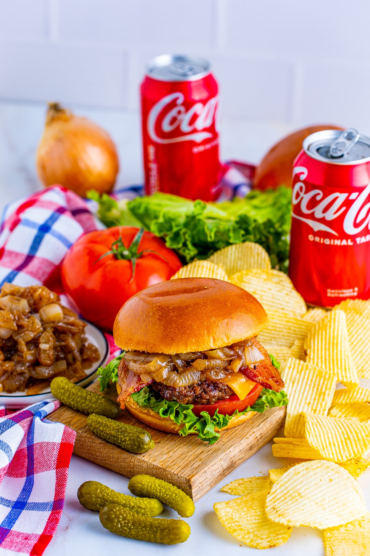 Bacon Cheeseburgers with Coca-Cola Onions on wooden plate with soda cans in back with onions and chips.