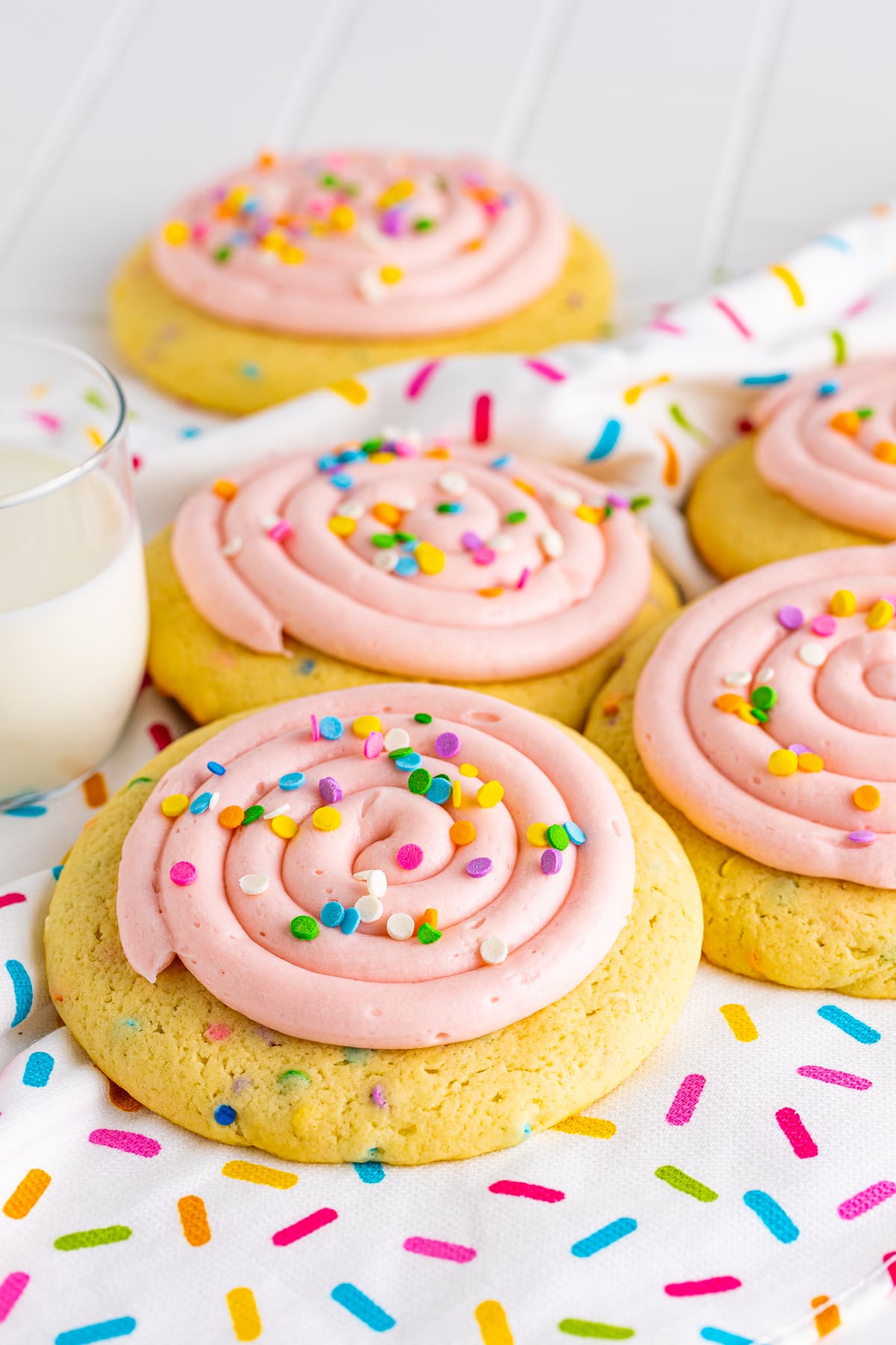 Lined up Confetti Cake Cookies on sprinkled apron with glass of milk.