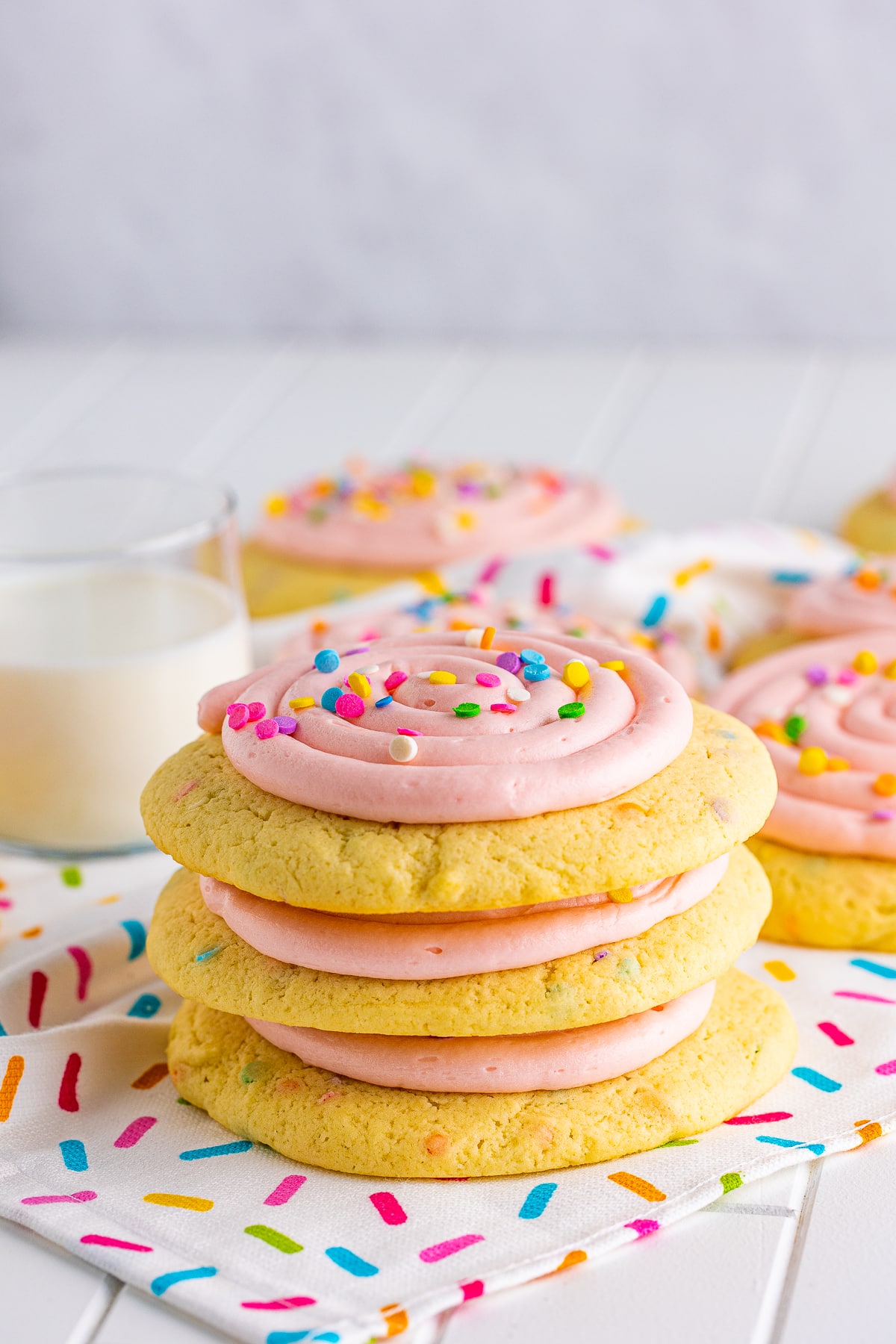 Three stacked Confetti Cake Cookies on linen with glass of milk behind it.