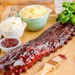 A rack of saucy smoked baby back ribs sliced on a wooden board with sides.