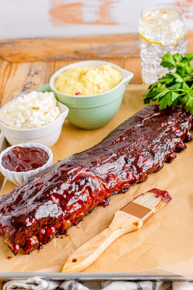 A rack of saucy smoked baby back ribs sliced on a wooden board with sides.