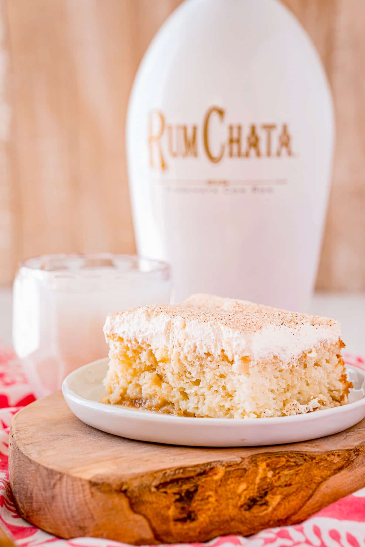 Slice of RumChata Poke Cake on white plate with glass of milk and alcohol in background.