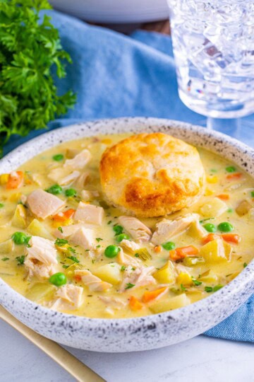 A white bowl of chicken pot pie soup topped with a flaky biscuit, with peas, corn, and shredded chicken in a creamy broth.