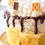 A tall s'mores cake on a cake stand with a chocolate ganache drip, toasted marshmallows, graham crackers, and chocolate squares on top.