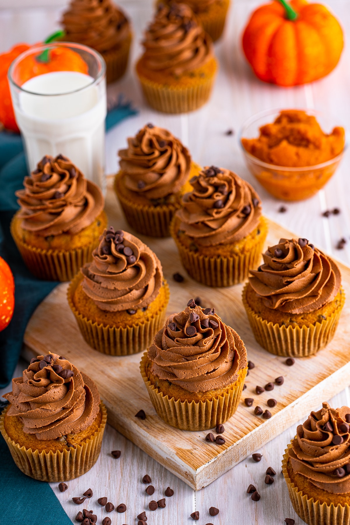 Chocolate Chip Pumpkin Cupcakes on wooden board with milk, chocolate chips and pumpkins in background.