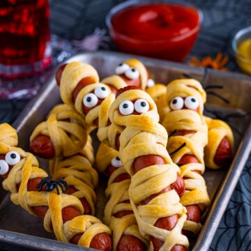 Mummy Dogs on metal tray stacked with ketchup and drink in background.