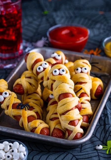 Mummy Dogs on metal tray stacked with ketchup and drink in background.