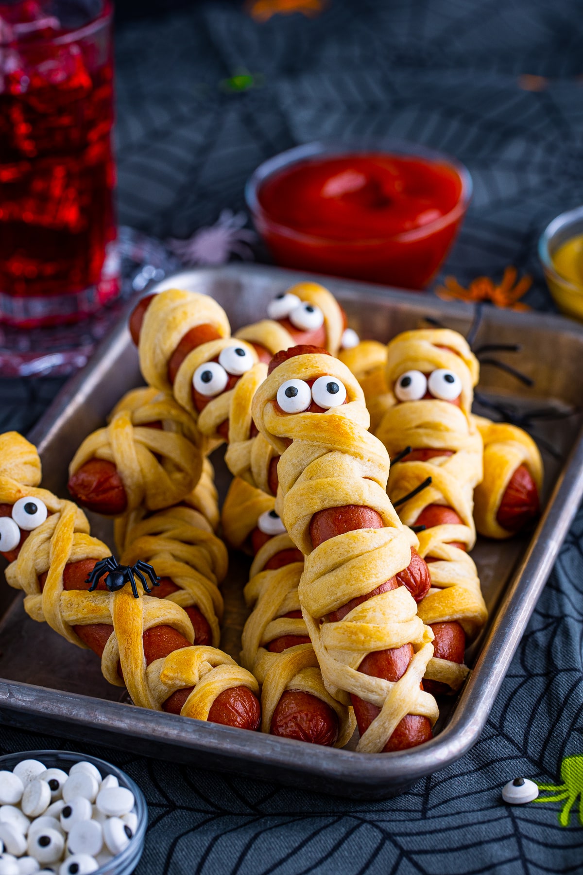 Mummy Dogs on metal tray stacked with ketchup and drink in background.