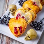 Heart shaped strawberry hand pies with red filling bite marks lined up on a white platter with candy skulls.