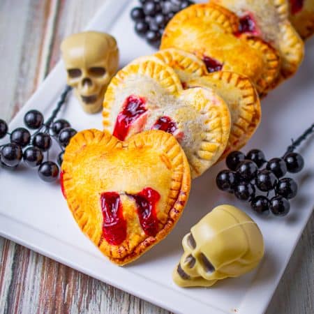 Heart shaped strawberry hand pies with red filling bite marks lined up on a white platter with candy skulls.