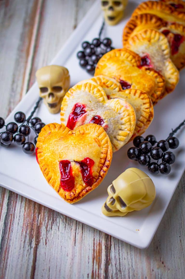 Heart shaped strawberry hand pies with red filling bite marks lined up on a white platter with candy skulls.