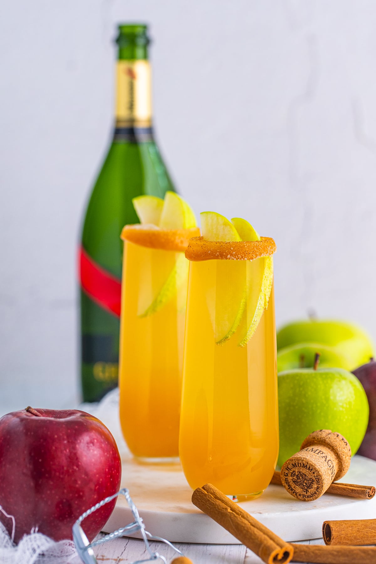 Two Apple Cider Mimosas on marble platter with champagne in background with apples and cinnamon around it.