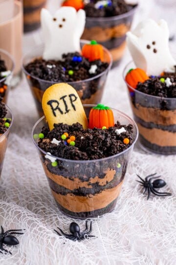 Halloween dirt cups topped with RIP Milano tombstones, a ghost Peep, and candy pumpkins on a spooky table.