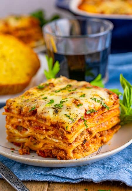 Slice of Homemade Lasagna on plate with drink and garlic toast in background.