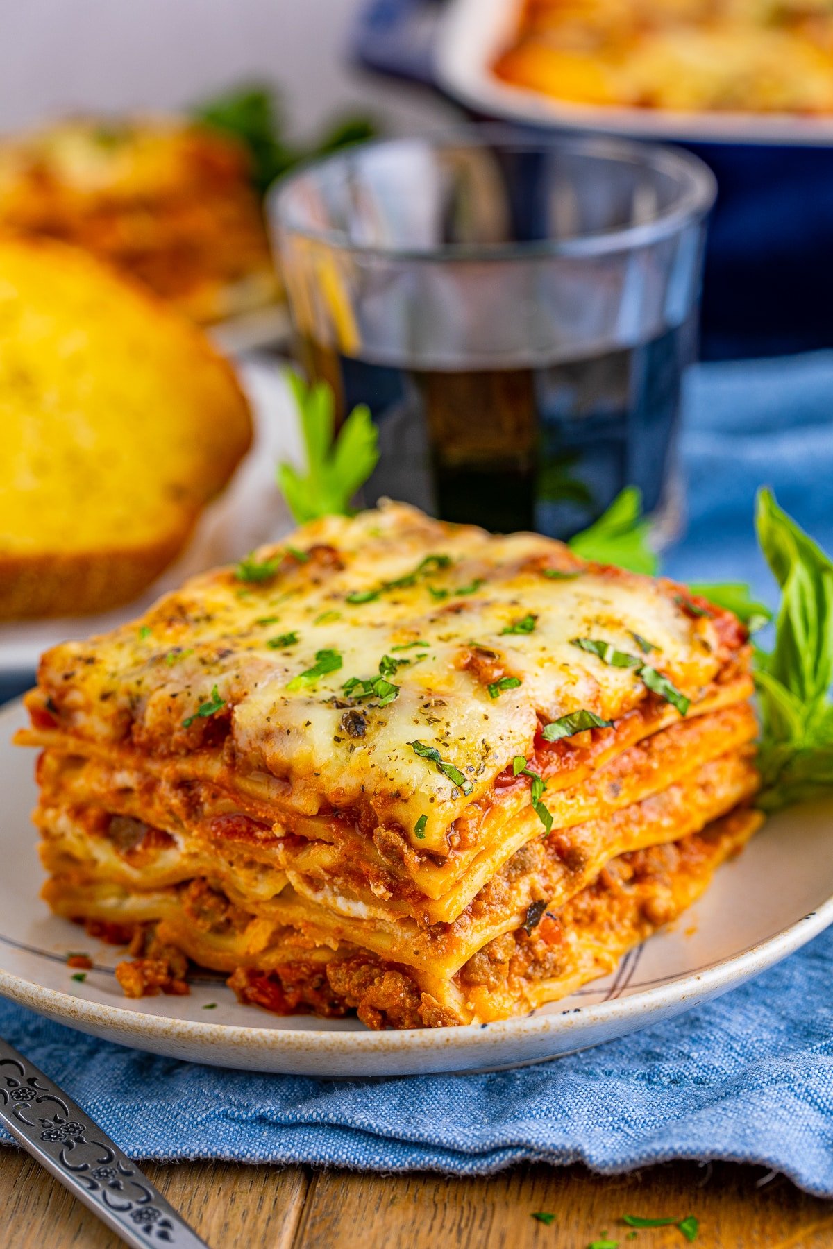 Slice of Homemade Lasagna on plate with drink and garlic toast in background.