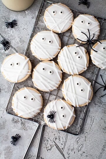 Mummy cookies with royal icing wrapping and candy eyes on a wire rack with plastic spiders.