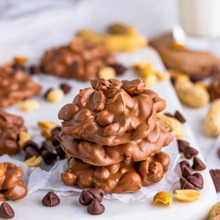 Three stacked Slow Cooker Peanut Clusters with chocolate chips and peanuts around them.
