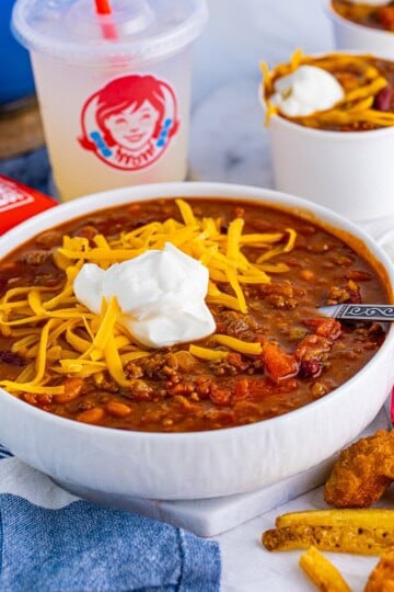 A bowl of Wendys chili topped with shredded cheddar cheese and sour cream next to a Wendys cup