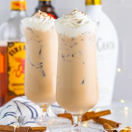 Cinnamon Roll Cocktail finished in two glasses with alcohol in background and cinnamon sticks around glasses.