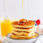 Syrup being poured over the stack of pancakes on white plate.