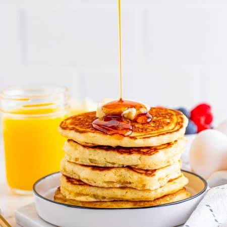 Syrup being poured over the stack of pancakes on white plate.