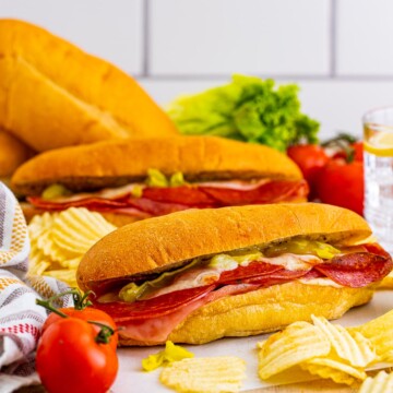 Two of the Italian Subs on parchment paper with chips surrounding it and a tomato in front.