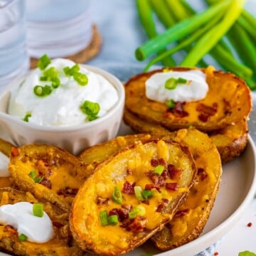 Finished Loaded Potato Skins on white plate with toppings and sour cream for dipping on side.