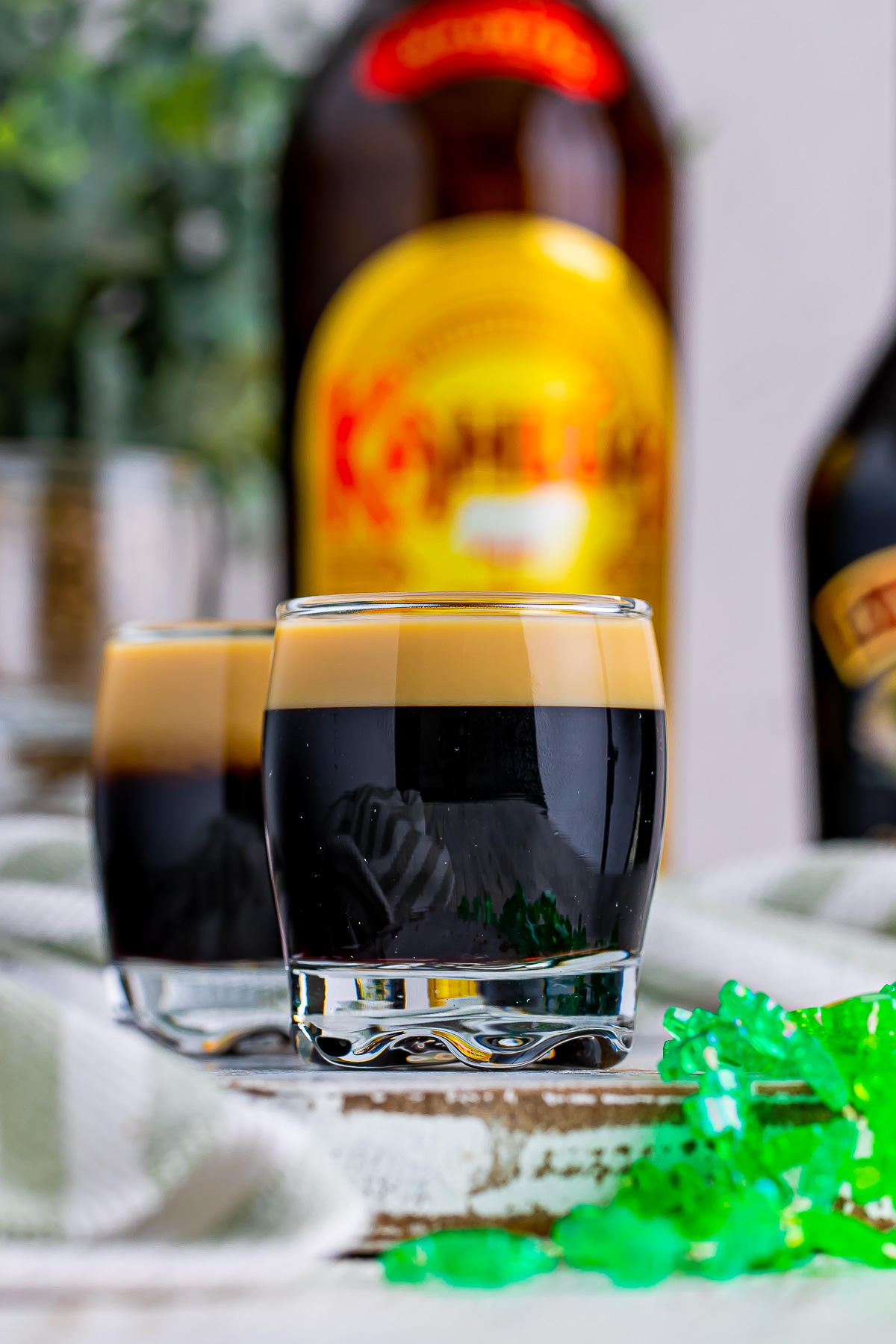 Close-up straight on image of Baby Guinness Shots with festive green clover necklace. Bottle of Kahlua in the background on a chipped wood serving board.