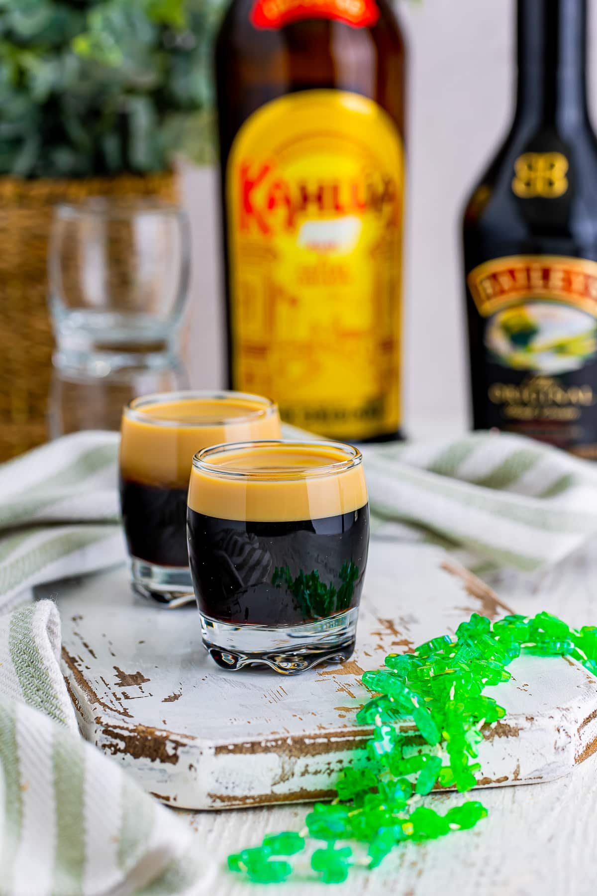 Baby Guinness Shots are on a white chipped wooden serving board with green festive necklace. Alcohol bottles in the background with striped green linen.