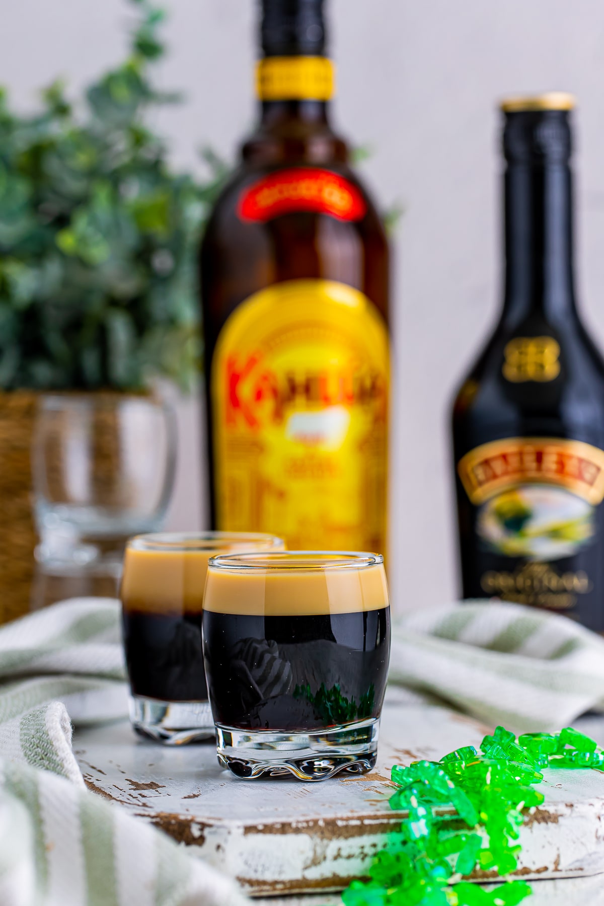Straight on image of Baby Guinness Shots on a white wooden chipped serving board, festive green necklace in foreground, alcohol bottles in background. Plant in a thicker holder in background, green striped linen.
