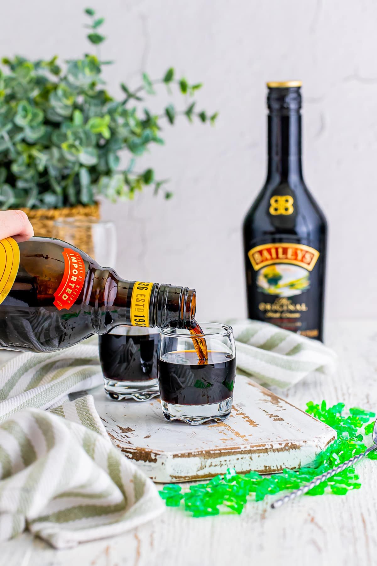 Kahlua being poured into a shot glass for Baby Guinness Shots on a white chipped wooden serving board, green festive necklace. Plant in wicker holder in background