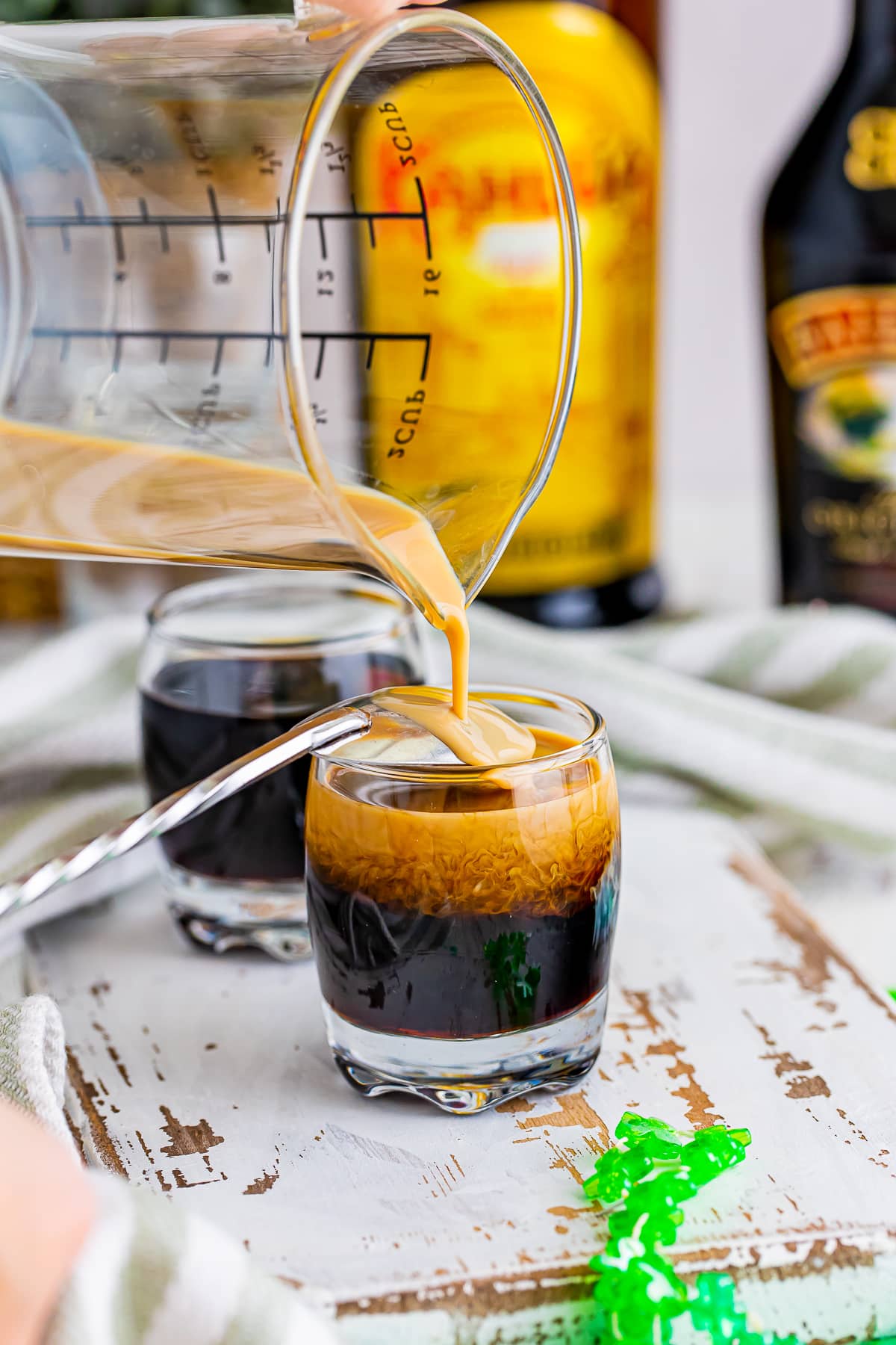 Showing how to invert spoon over the top of the Baby Guinness Shots and slowly pour the Bailey's Irish Cream on top. Shown on a white chipped wooden serving board