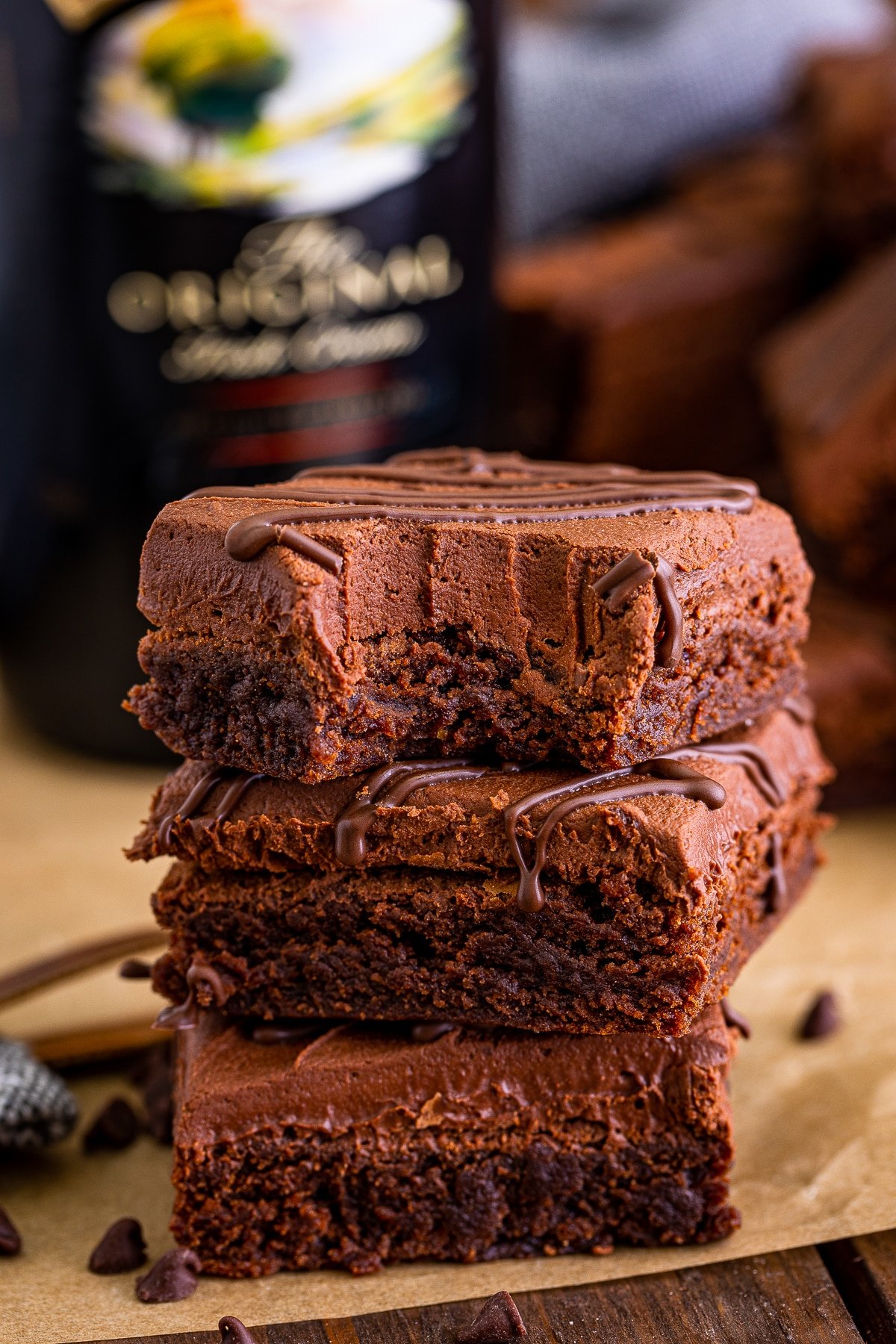 A 3 high stack of Bailey's Brownies with a bite taken out on top on brown parchment paper. Alcohol bottle in the background