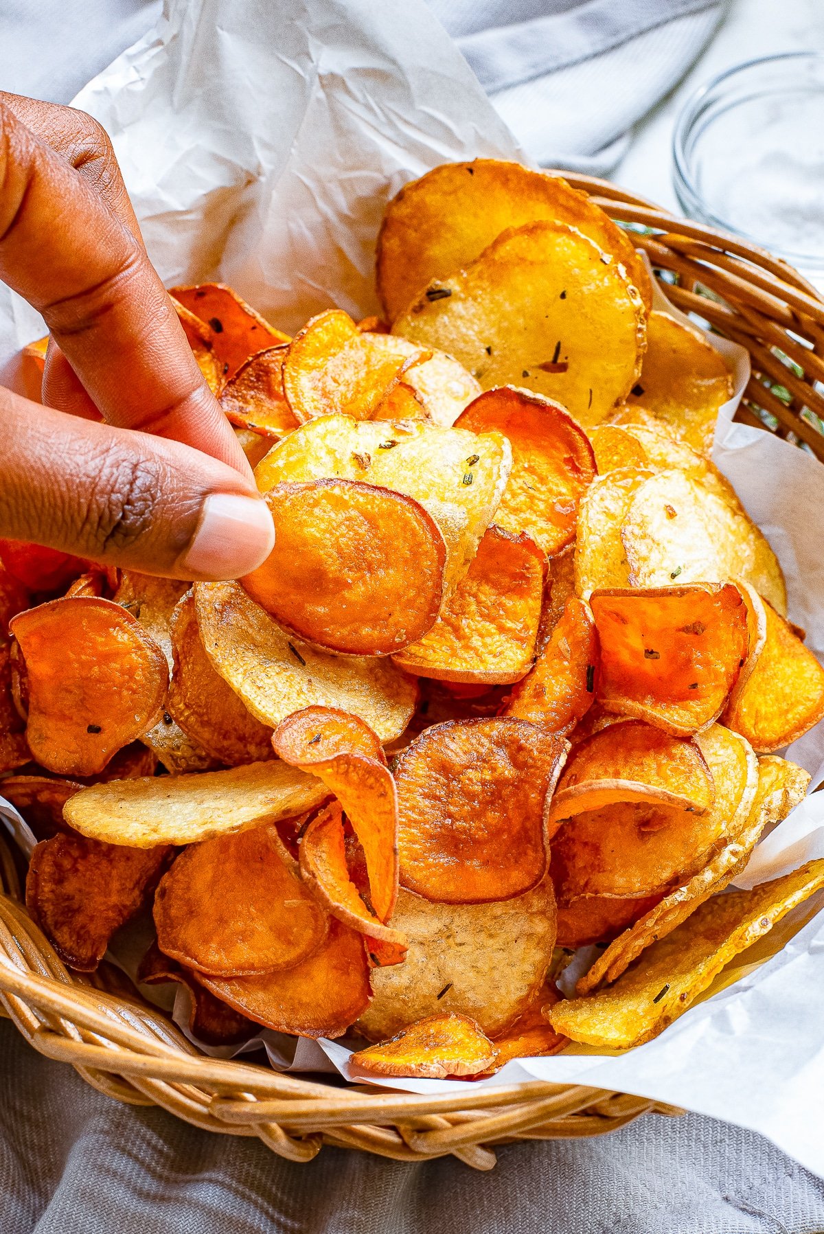Baked potato chips in a wooden bowl with parchment paper and a hand grabbing one.